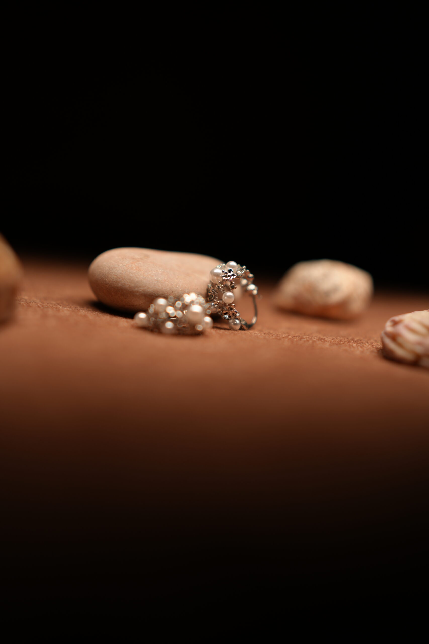A pair of silver-tone hoop earrings encrusted with faux pearl clusters and pave crystal accents, resting against a smooth beige pebble on a warm brown fabric surface surrounded by seashells.