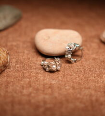 A pair of silver-tone hoop earrings encrusted with faux pearl clusters and pave crystal accents, resting against a smooth beige pebble on a warm brown fabric surface surrounded by seashells.