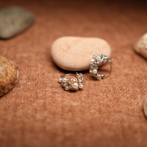 A pair of silver-tone hoop earrings encrusted with faux pearl clusters and pave crystal accents, resting against a smooth beige pebble on a warm brown fabric surface surrounded by seashells.