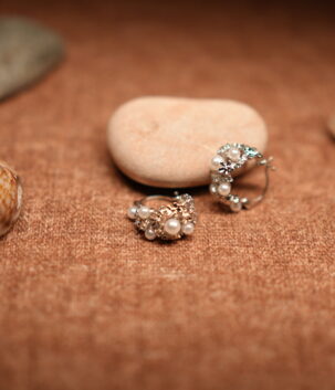 A pair of silver-tone hoop earrings encrusted with faux pearl clusters and pave crystal accents, resting against a smooth beige pebble on a warm brown fabric surface surrounded by seashells.