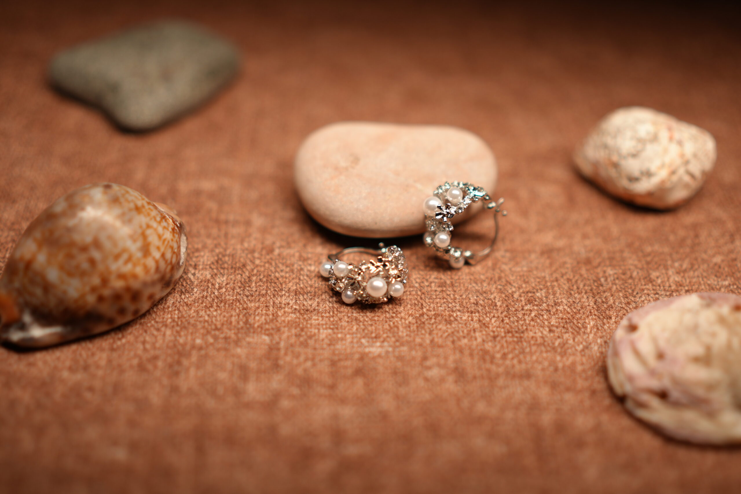A pair of silver-tone hoop earrings encrusted with faux pearl clusters and pave crystal accents, resting against a smooth beige pebble on a warm brown fabric surface surrounded by seashells.