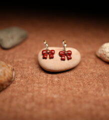 A pair of red glossy enamel bow drop earrings hanging from small crystal-studded gold-tone hoops, displayed on a smooth beige pebble against a warm brown fabric background with seashells.