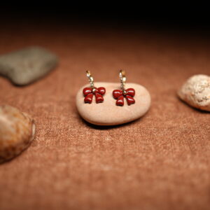 A pair of red glossy enamel bow drop earrings hanging from small crystal-studded gold-tone hoops, displayed on a smooth beige pebble against a warm brown fabric background with seashells.