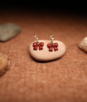 A pair of red glossy enamel bow drop earrings hanging from small crystal-studded gold-tone hoops, displayed on a smooth beige pebble against a warm brown fabric background with seashells.