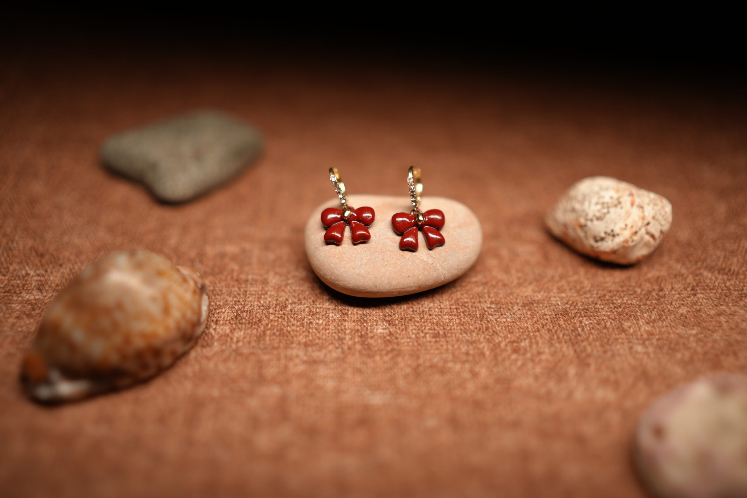 A pair of red glossy enamel bow drop earrings hanging from small crystal-studded gold-tone hoops, displayed on a smooth beige pebble against a warm brown fabric background with seashells.