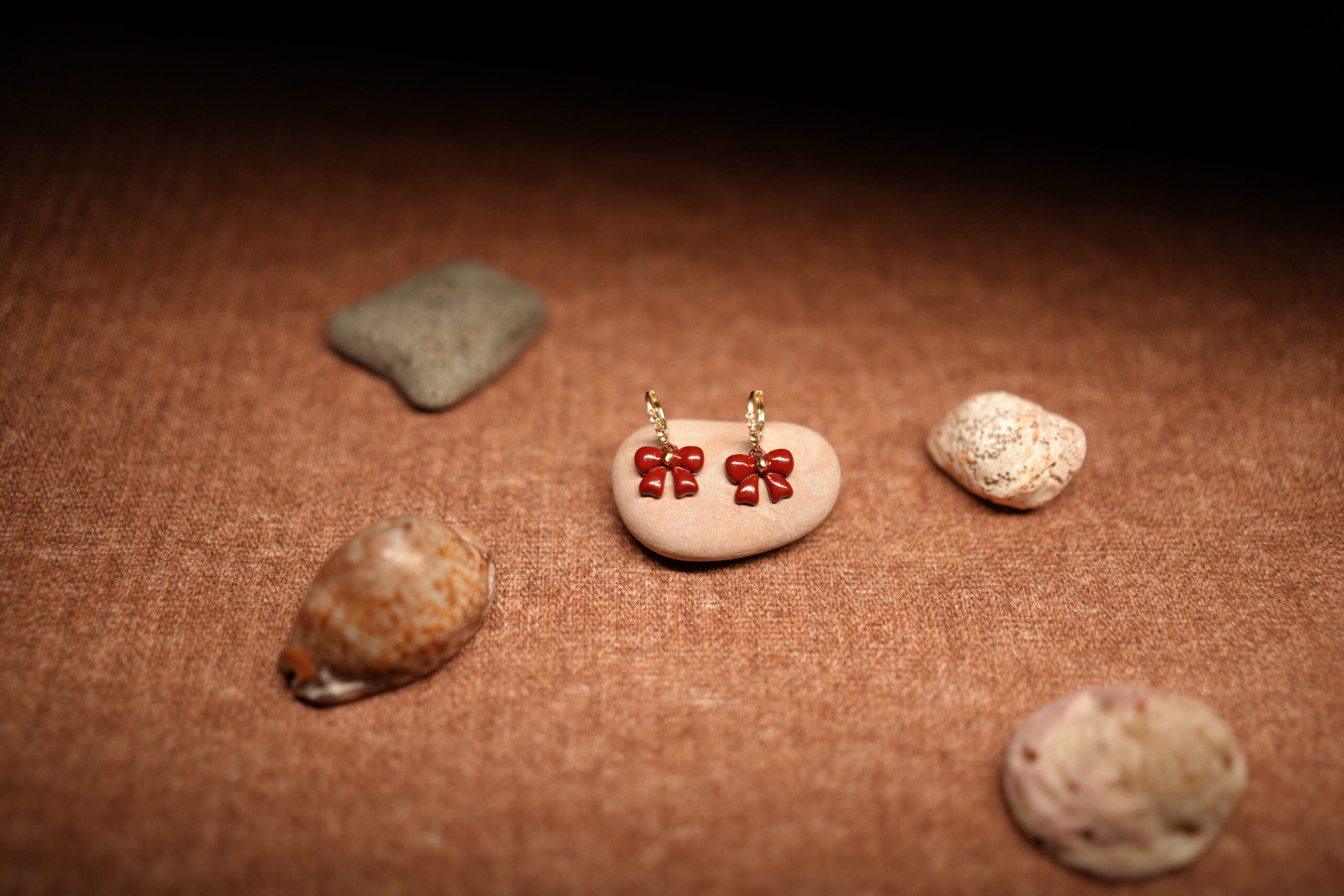 A pair of red glossy enamel bow drop earrings hanging from small crystal-studded gold-tone hoops, displayed on a smooth beige pebble against a warm brown fabric background with seashells.