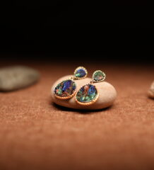 A pair of gold-tone double-drop earrings featuring a small teardrop and large oval abalone shell cabochon in beaded frames, resting on a smooth beige pebble against a warm brown fabric background with seashells.