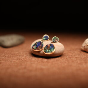 A pair of gold-tone double-drop earrings featuring a small teardrop and large oval abalone shell cabochon in beaded frames, resting on a smooth beige pebble against a warm brown fabric background with seashells.