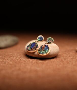 A pair of gold-tone double-drop earrings featuring a small teardrop and large oval abalone shell cabochon in beaded frames, resting on a smooth beige pebble against a warm brown fabric background with seashells.