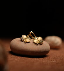 A pair of gold-tone flower stud earrings with cream moonstone-effect petals and pave crystal accents, resting on a smooth terracotta-toned pebble against a dark background.