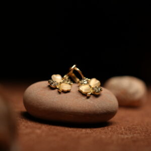A pair of gold-tone flower stud earrings with cream moonstone-effect petals and pave crystal accents, resting on a smooth terracotta-toned pebble against a dark background.