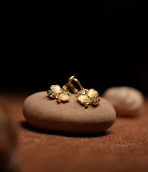A pair of gold-tone flower stud earrings with cream moonstone-effect petals and pave crystal accents, resting on a smooth terracotta-toned pebble against a dark background.