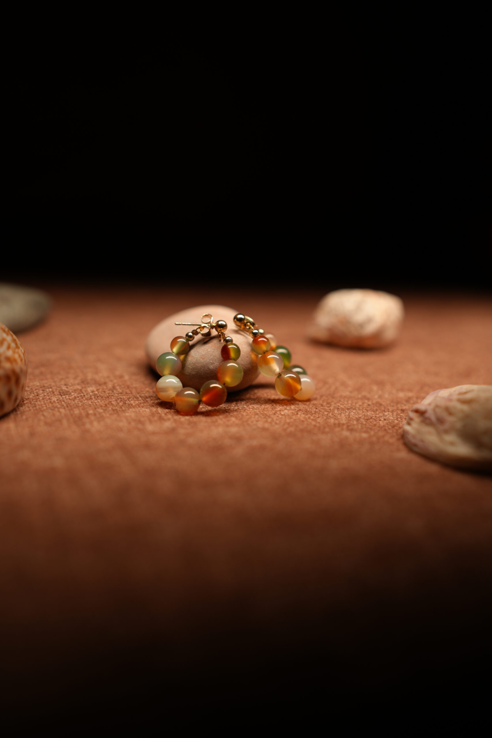 A pair of gold-tone hoop earrings strung with multicolour agate-effect round beads in terracotta, sage green, amber, and ivory tones with gold ball spacers, resting on a smooth beige pebble against a warm brown fabric background.