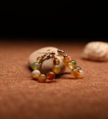 A pair of gold-tone hoop earrings strung with multicolour agate-effect round beads in terracotta, sage green, amber, and ivory tones with gold ball spacers, resting on a smooth beige pebble against a warm brown fabric background.