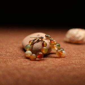 A pair of gold-tone hoop earrings strung with multicolour agate-effect round beads in terracotta, sage green, amber, and ivory tones with gold ball spacers, resting on a smooth beige pebble against a warm brown fabric background.