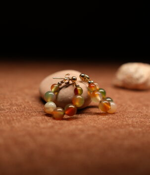 A pair of gold-tone hoop earrings strung with multicolour agate-effect round beads in terracotta, sage green, amber, and ivory tones with gold ball spacers, resting on a smooth beige pebble against a warm brown fabric background.