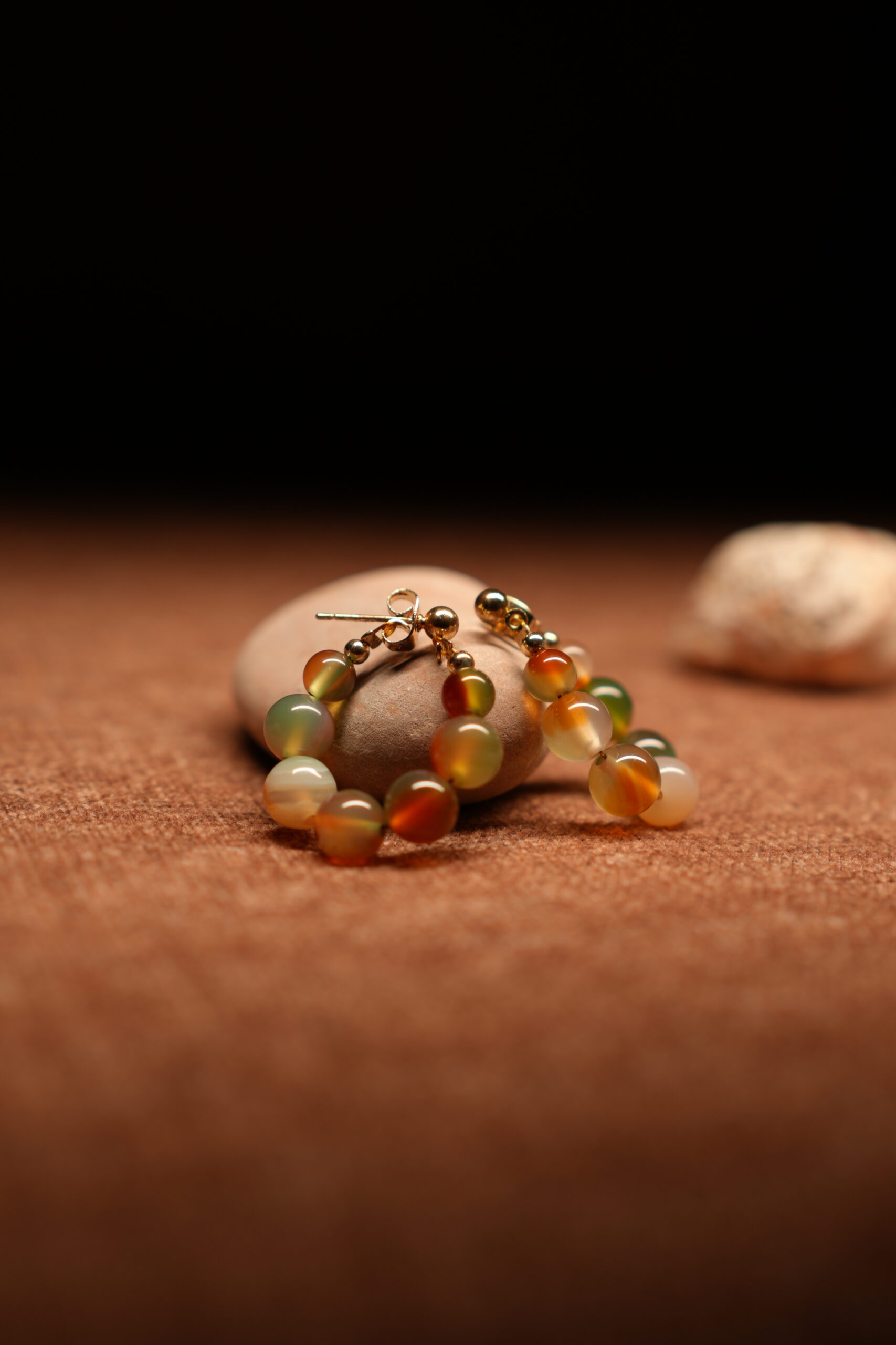 A pair of gold-tone hoop earrings strung with multicolour agate-effect round beads in terracotta, sage green, amber, and ivory tones with gold ball spacers, resting on a smooth beige pebble against a warm brown fabric background.