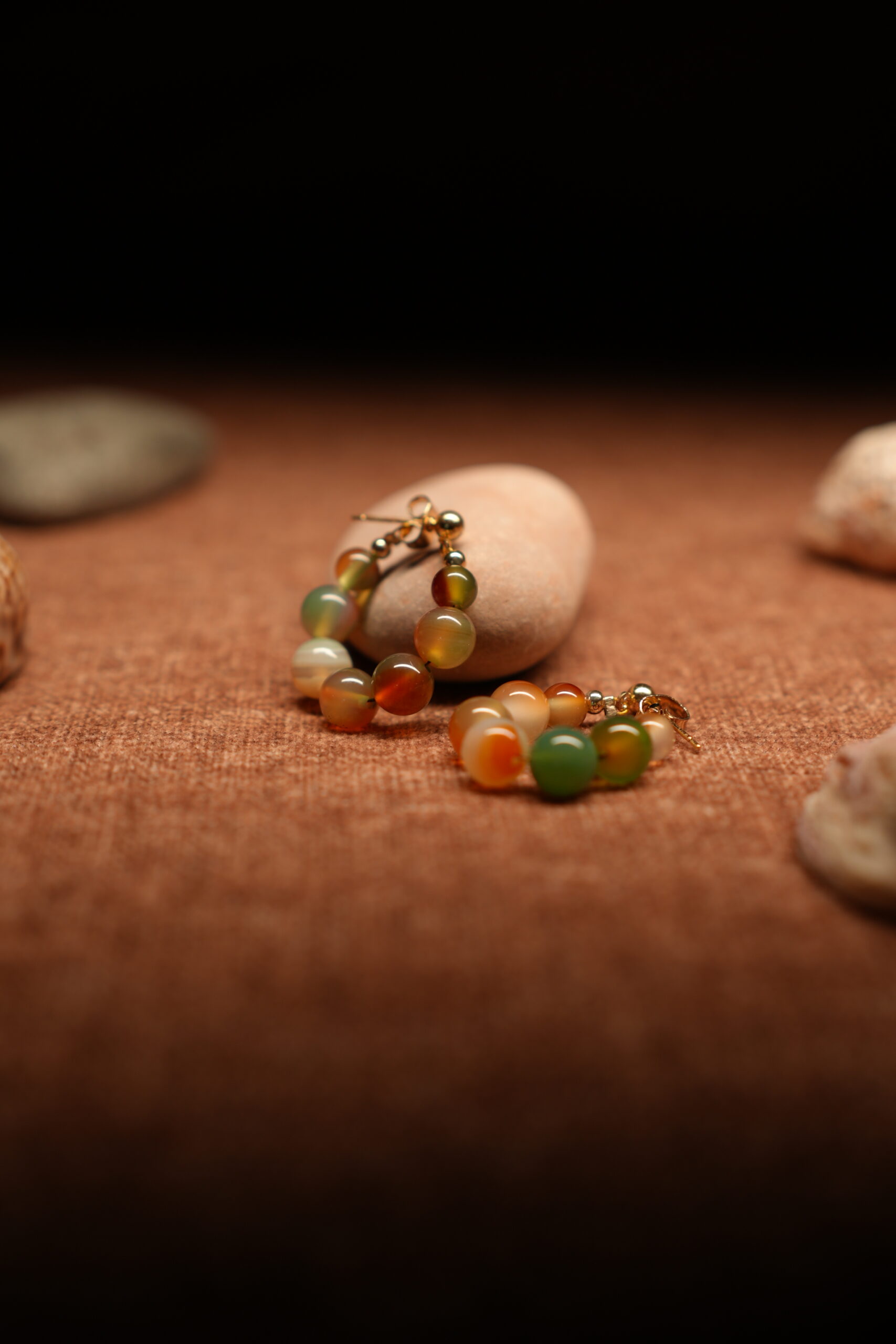A pair of gold-tone hoop earrings strung with multicolour agate-effect round beads in terracotta, sage green, amber, and ivory tones with gold ball spacers, resting on a smooth beige pebble against a warm brown fabric background.