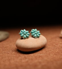 A pair of turquoise teardrop bead cluster flower stud earrings in a gold-tone setting, displayed on a smooth beige pebble against a warm brown fabric background with seashells and stones.