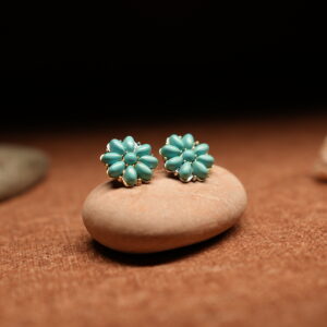 A pair of turquoise teardrop bead cluster flower stud earrings in a gold-tone setting, displayed on a smooth beige pebble against a warm brown fabric background with seashells and stones.