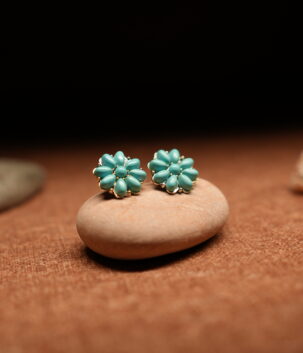 A pair of turquoise teardrop bead cluster flower stud earrings in a gold-tone setting, displayed on a smooth beige pebble against a warm brown fabric background with seashells and stones.