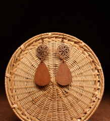 A pair of copper-tone filigree mandala disc and natural walnut wood teardrop drop earrings on French hooks, displayed against a round woven rattan disc on a warm brown background.
