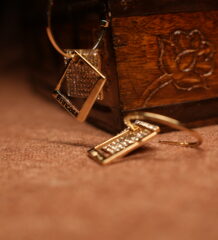 A pair of gold-tone open hoop earrings with a pave crystal diamond-shaped top and a channel-set crystal rectangular bar pendant, displayed against a carved wooden jewellery box on a warm brown surface.