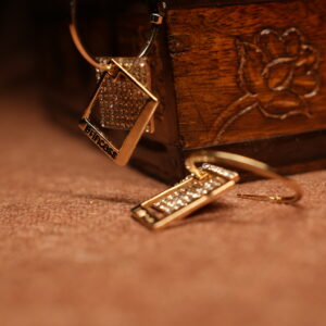 A pair of gold-tone open hoop earrings with a pave crystal diamond-shaped top and a channel-set crystal rectangular bar pendant, displayed against a carved wooden jewellery box on a warm brown surface.
