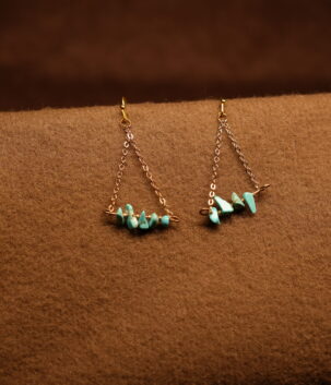 A pair of gold-tone chain drop earrings with a triangular silhouette, featuring a row of raw turquoise chip stones on a horizontal bar, displayed on a warm brown fabric surface.