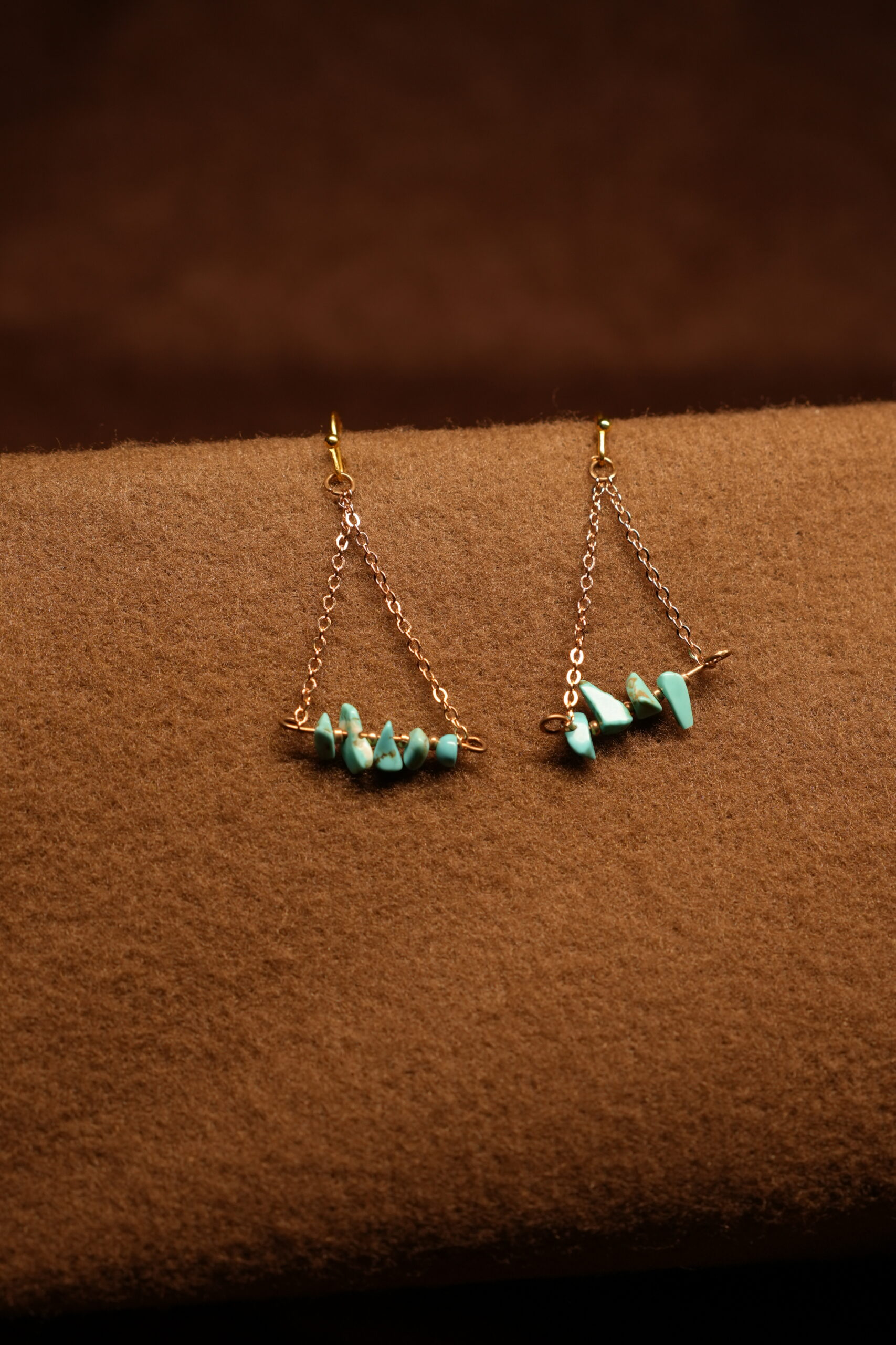A pair of gold-tone chain drop earrings with a triangular silhouette, featuring a row of raw turquoise chip stones on a horizontal bar, displayed on a warm brown fabric surface.