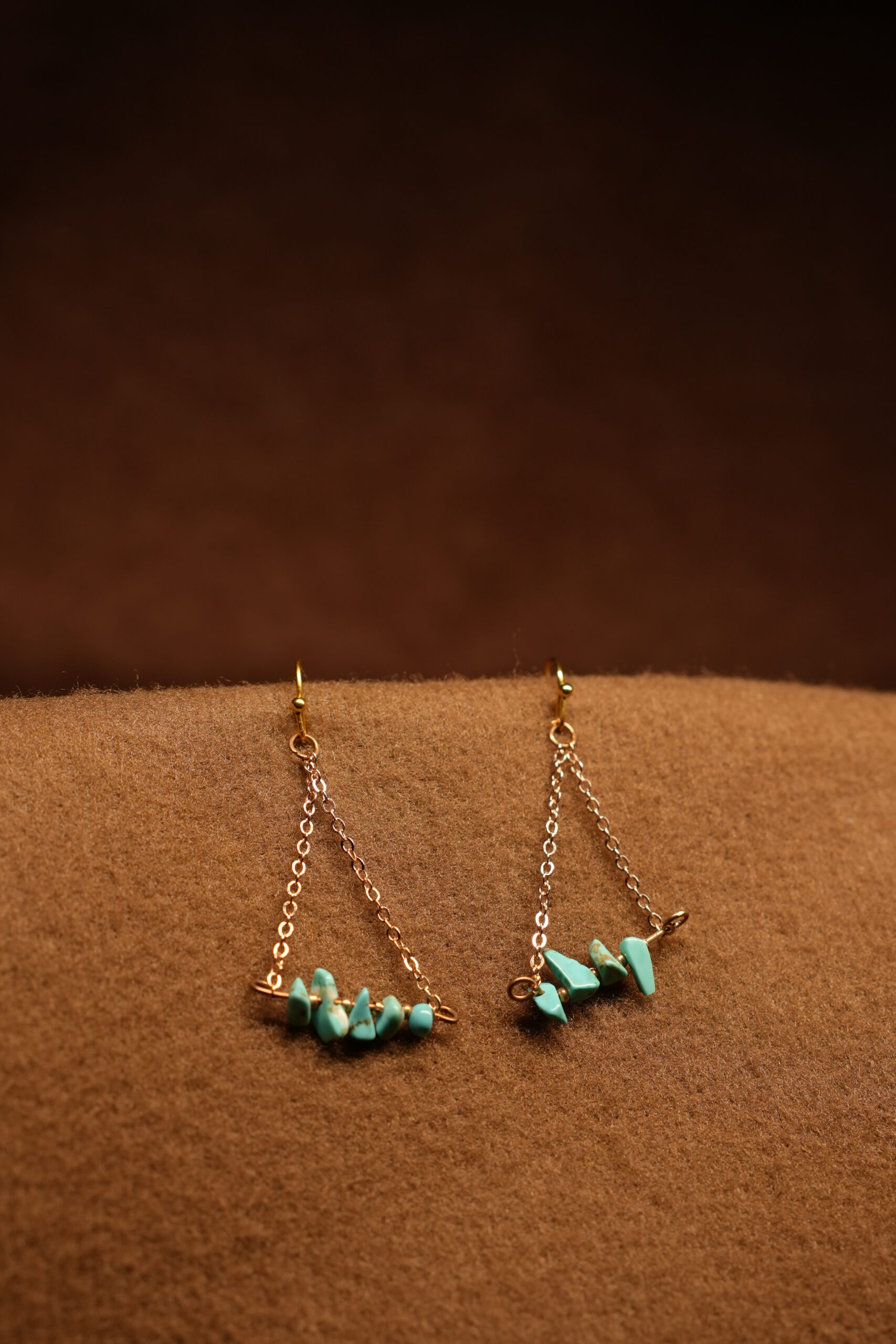 A pair of gold-tone chain drop earrings with a triangular silhouette, featuring a row of raw turquoise chip stones on a horizontal bar, displayed on a warm brown fabric surface.