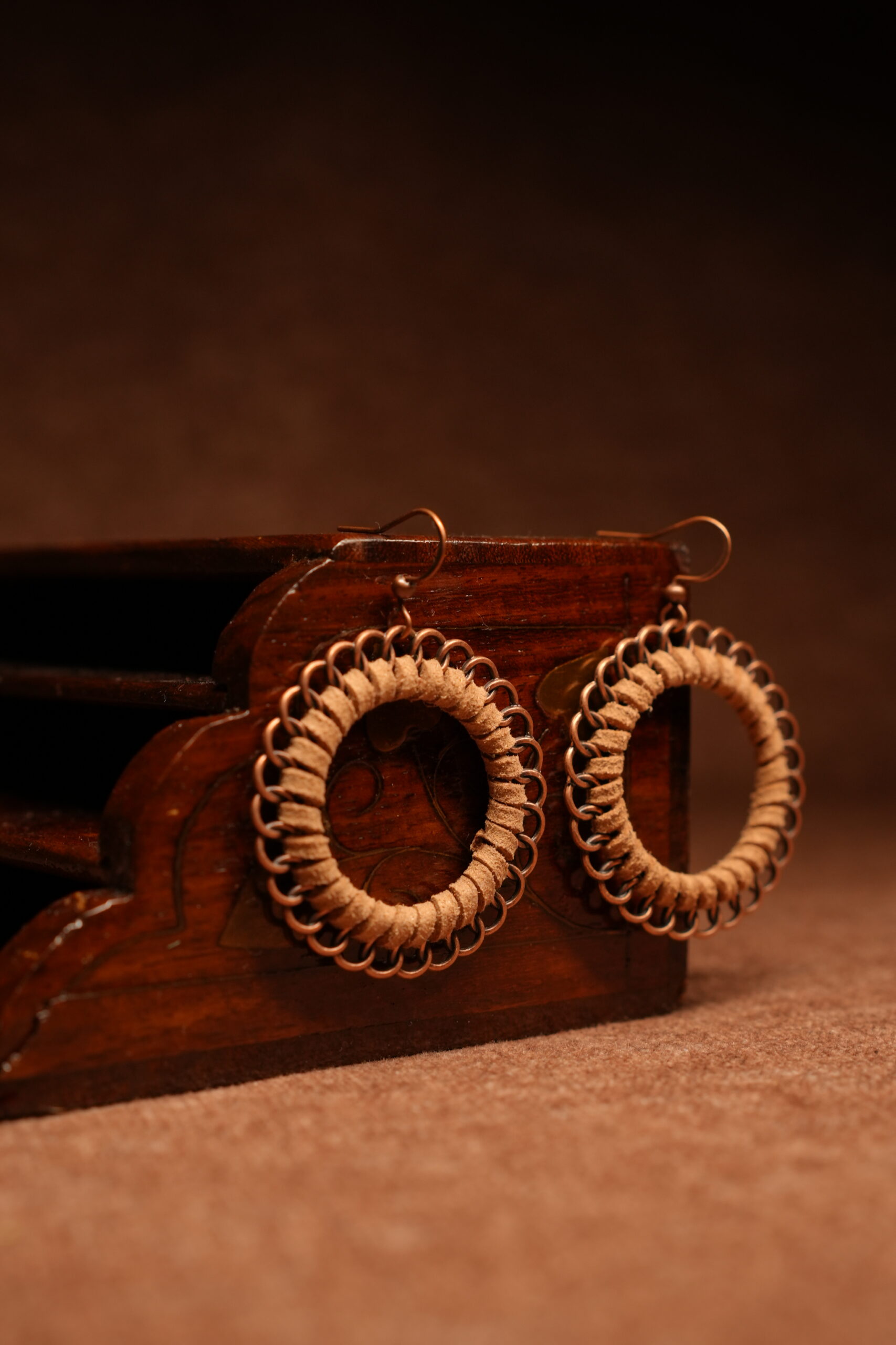 A pair of large circular hoop earrings made with tan faux suede cord woven through copper-tone chainmail jump rings, displayed against a carved dark wooden jewellery box on a warm brown surface.