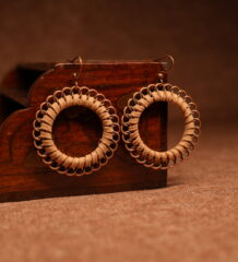 A pair of large circular hoop earrings made with tan faux suede cord woven through copper-tone chainmail jump rings, displayed against a carved dark wooden jewellery box on a warm brown surface.