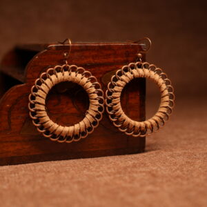 A pair of large circular hoop earrings made with tan faux suede cord woven through copper-tone chainmail jump rings, displayed against a carved dark wooden jewellery box on a warm brown surface.