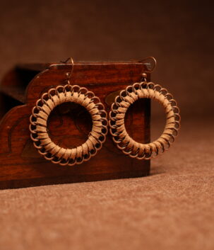 A pair of large circular hoop earrings made with tan faux suede cord woven through copper-tone chainmail jump rings, displayed against a carved dark wooden jewellery box on a warm brown surface.
