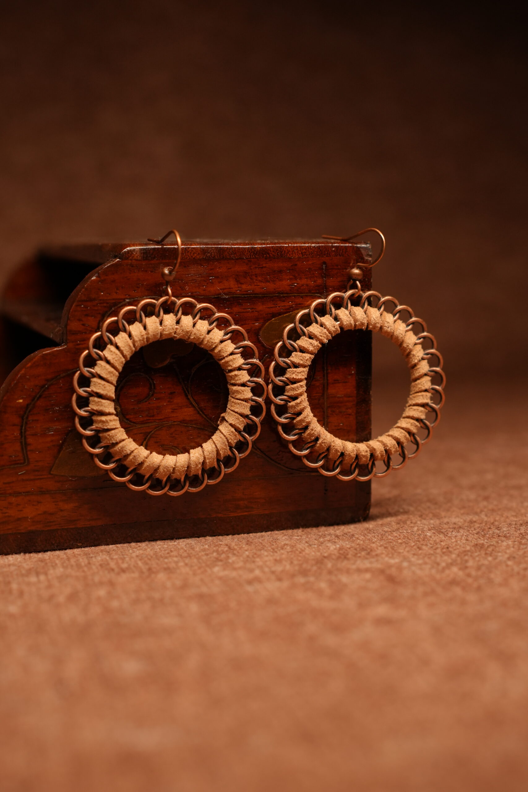 A pair of large circular hoop earrings made with tan faux suede cord woven through copper-tone chainmail jump rings, displayed against a carved dark wooden jewellery box on a warm brown surface.
