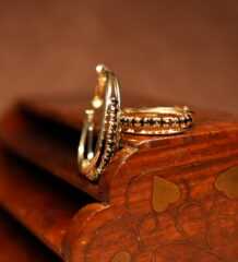 A pair of gold-tone hoop earrings with a continuous row of prong-set black round crystals along the inner edge, displayed upright against a carved dark wooden jewellery box on a warm brown surface.