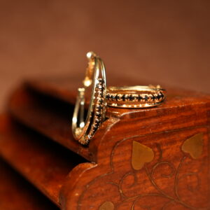 A pair of gold-tone hoop earrings with a continuous row of prong-set black round crystals along the inner edge, displayed upright against a carved dark wooden jewellery box on a warm brown surface.