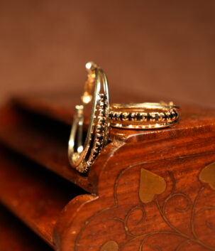A pair of gold-tone hoop earrings with a continuous row of prong-set black round crystals along the inner edge, displayed upright against a carved dark wooden jewellery box on a warm brown surface.
