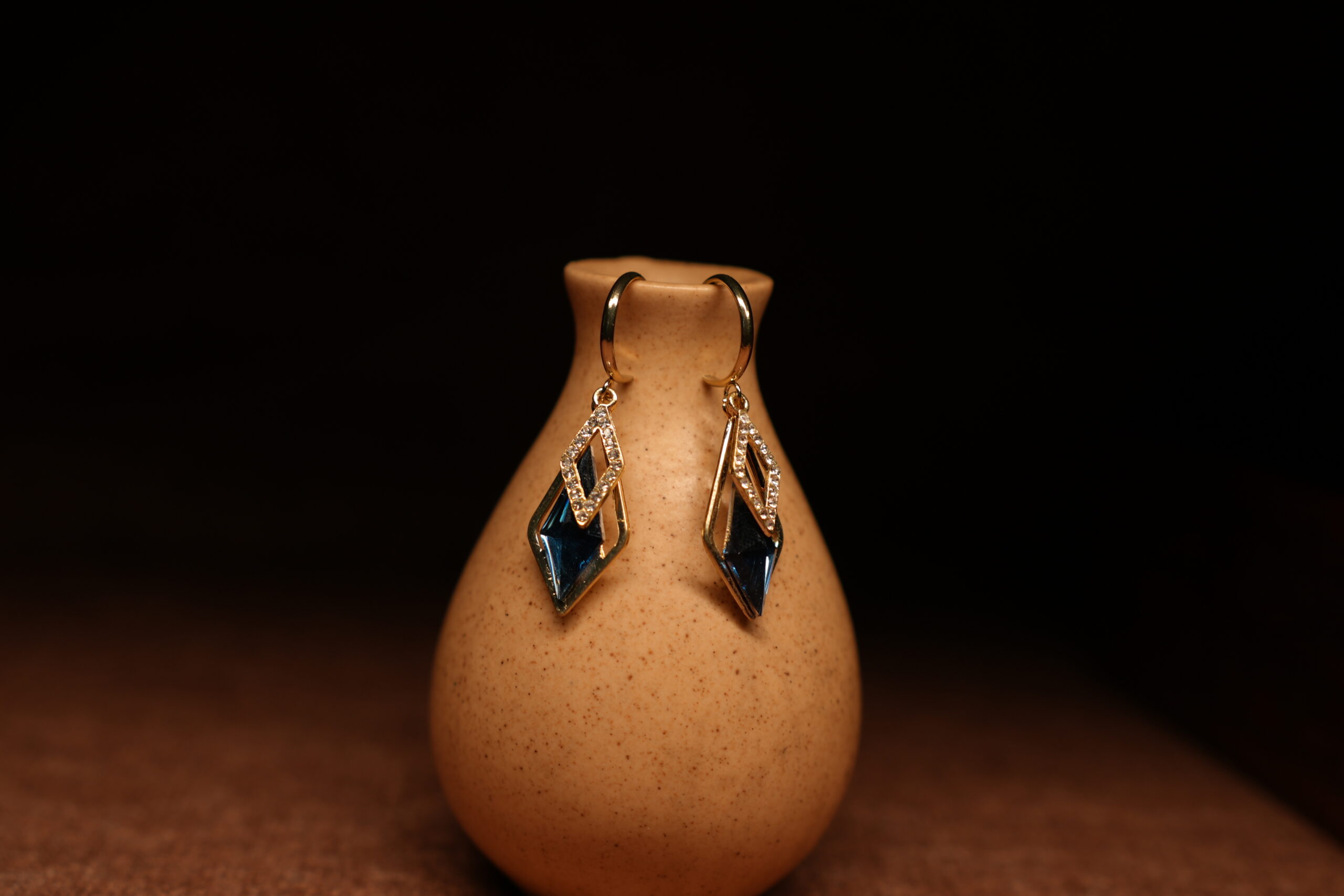 A pair of gold-tone mini hoop earrings with a pave crystal diamond-shaped frame and a large deep sapphire blue faceted kite crystal pendant, displayed on a speckled beige ceramic vase against a dark background.