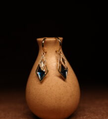 A pair of gold-tone mini hoop earrings with a pave crystal diamond-shaped frame and a large deep sapphire blue faceted kite crystal pendant, displayed on a speckled beige ceramic vase against a dark background.