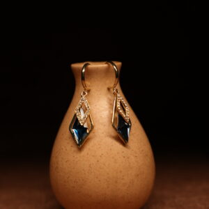 A pair of gold-tone mini hoop earrings with a pave crystal diamond-shaped frame and a large deep sapphire blue faceted kite crystal pendant, displayed on a speckled beige ceramic vase against a dark background.