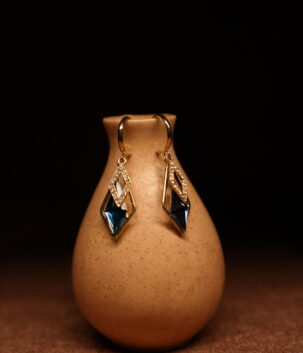 A pair of gold-tone mini hoop earrings with a pave crystal diamond-shaped frame and a large deep sapphire blue faceted kite crystal pendant, displayed on a speckled beige ceramic vase against a dark background.