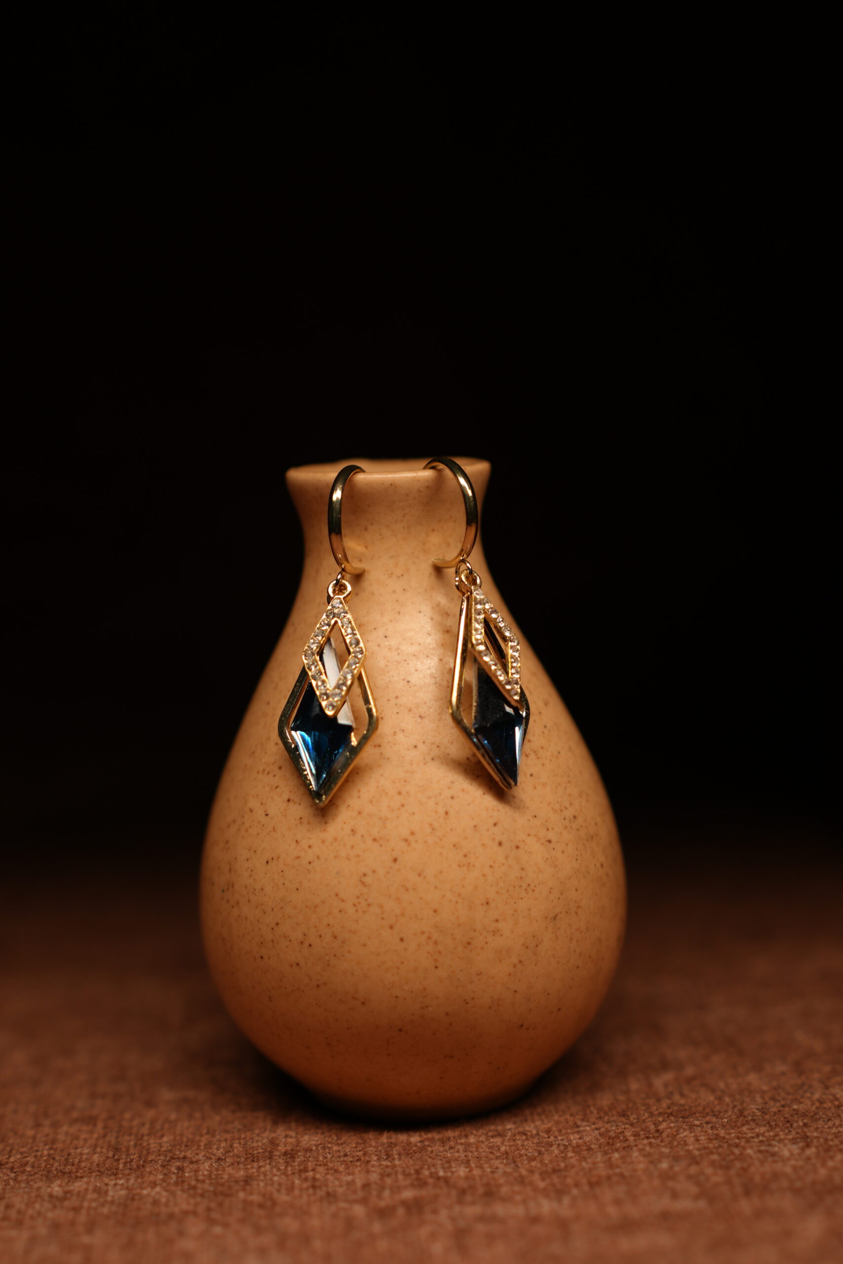 A pair of gold-tone mini hoop earrings with a pave crystal diamond-shaped frame and a large deep sapphire blue faceted kite crystal pendant, displayed on a speckled beige ceramic vase against a dark background.