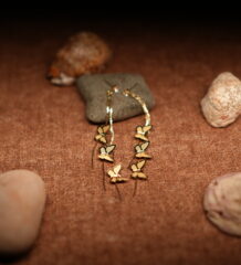 A pair of long gold-tone chain drop earrings with three graduating butterfly charms in descending sizes, displayed against a dark grey stone on a warm brown fabric surface with seashells.
