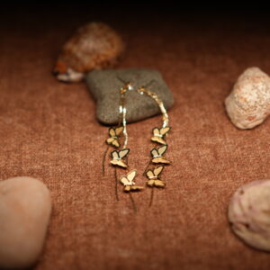 A pair of long gold-tone chain drop earrings with three graduating butterfly charms in descending sizes, displayed against a dark grey stone on a warm brown fabric surface with seashells.