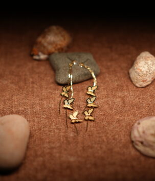 A pair of long gold-tone chain drop earrings with three graduating butterfly charms in descending sizes, displayed against a dark grey stone on a warm brown fabric surface with seashells.