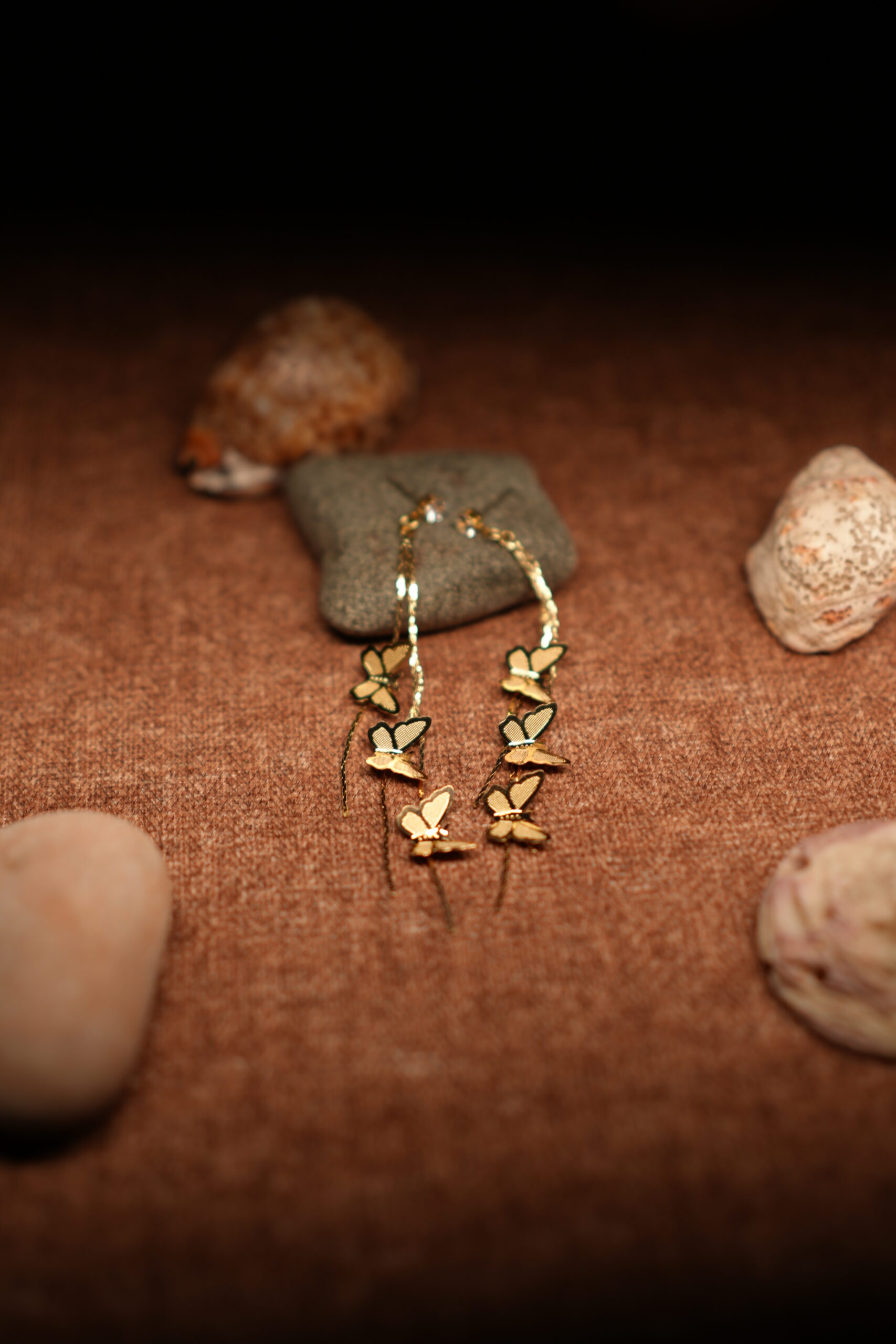 A pair of long gold-tone chain drop earrings with three graduating butterfly charms in descending sizes, displayed against a dark grey stone on a warm brown fabric surface with seashells.