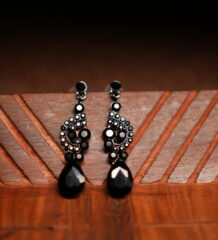 A pair of gunmetal-tone chandelier drop earrings with an ornate black crystal cluster body and a large faceted black teardrop pendant, displayed on a geometric carved wooden surface against a dark background.