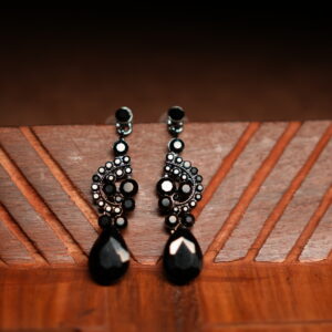 A pair of gunmetal-tone chandelier drop earrings with an ornate black crystal cluster body and a large faceted black teardrop pendant, displayed on a geometric carved wooden surface against a dark background.